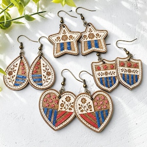 May include: A collection of wooden earrings in various shapes: teardrop, star, shield, and heart. Each earring features a patriotic design with red, white, and blue elements, floral accents, and brass-colored ear wires. The earrings are displayed on a white surface.