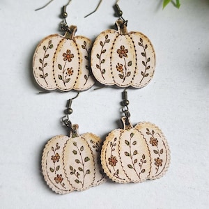 Puede incluir: Dos pares de pendientes colgantes con formas de calabaza de madera en color crema. Cada calabaza está decorada con dibujos de líneas quemadas de pequeñas flores naranjas y enredaderas verdes. Cuelgan de ganchos y anillas de metal oscuro.