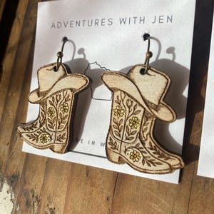 May include: Wooden cowboy boot earrings with floral details and a cowboy hat design. The earrings are hanging on a white card with the text "ADVENTURES WITH JEN". The earrings are made of light-colored wood.