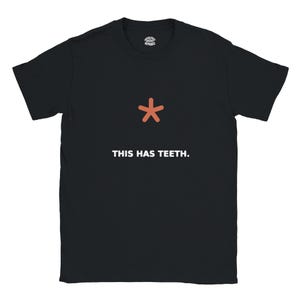 May include: A black short-sleeved t-shirt featuring a small orange asterisk symbol centered on the chest. Below the symbol, the text "THIS HAS TEETH." is printed in white capital letters. The shirt has a crew neckline and a standard fit.