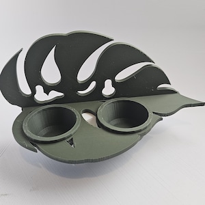 May include: A dark olive green wall-mounted shelf featuring a decorative cutout design shaped like a large monstera leaf. The shelf includes two circular holders integrated into the base, providing space for small items or containers.