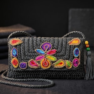 Op de afbeelding: Een donkergrijze gehaakte schoudertas met een rechthoekige klep versierd met levendige, veelkleurige geborduurde paisley- en bloemmotieven. De tas is voorzien van een bijpassende donkergrijze schouderband en een decoratieve kwast met kleine houten kralen.
