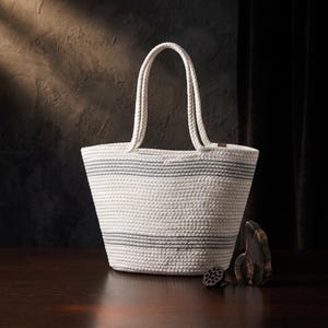 Op de afbeelding: Een witte tote bag gemaakt van opgerold touw met twee bijpassende gevlochten handvatten. De tas is voorzien van horizontale grijze strepen bij de boven- en onderrand. De textuur is uniform en geweven, wat een gestructureerde tas vormt.