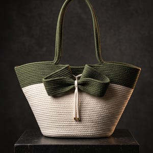 Op de afbeelding: Een tweekleurige tas gemaakt van gewikkeld touw, met een olijfgroen bovengedeelte en een crèmekleurig ondergedeelte. De tas is voorzien van twee olijfgroene touwhengsels en een grote decoratieve olijfgroene touwstrik in het midden.
