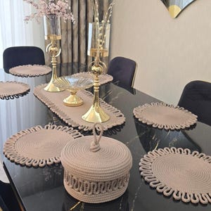 May include: A set of beige rope-textured table accessories including a round lidded container with a top loop handle, a long rectangular table runner, and several round placemats with decorative looped edges arranged on a black dining table.