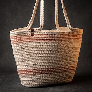 Op de afbeelding: Een draagtas gemaakt van opgerold gebroken wit touw met twee lange hengsels en twee horizontale strepen in een gedempte terracotta oranje kleur. Een klein bruin leren label met een subtiel logo is bevestigd aan de bovenrand van de tas.