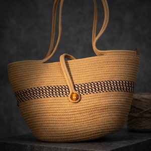 Op de afbeelding: Een beige touw-tote tas gemaakt van opgerold koord met twee lange hengsels. De tas heeft een horizontale band met een donkerbruin en beige geweven patroon in het midden, vastgezet met een enkele amberkleurige ronde knoopsluiting.
