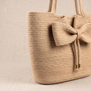 Op de afbeelding: Een beige tote bag gemaakt van gevlochten natuurvezeltouw. De tas is voorzien van een grote, opvallende strik op het voorpaneel, twee bijpassende touwhengsels en twee bungelende touwuiteinden met kleine, metalen goudkleurige cilindrische uiteinden.