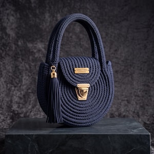 Op de afbeelding: Een marineblauwe ronde handtas gemaakt van dik koord met een gestructureerd handvat. De tas is voorzien van een goudkleurige metalen sluiting, een bijpassend rechthoekig goudkleurig label met de tekst DIMITRA en een lange marineblauwe kwast aan de zijkant.