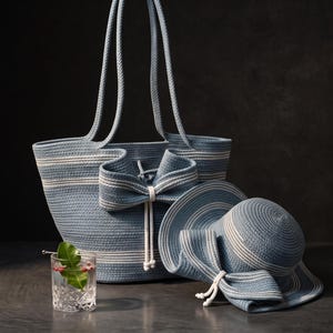 Op de afbeelding: Een blauw-witte gestreepte touw tas met een grote strik en een bijpassende hoed met brede rand. De tas heeft twee lange hengsels en een trekkoordsluiting. Een helder glas met een groen blaadje en rode bessen staat naast de voorwerpen.