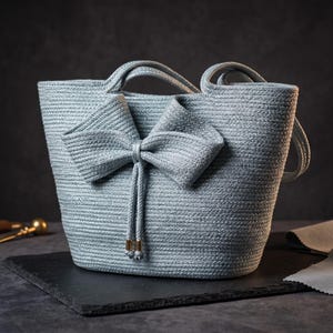 Op de afbeelding: Een lichtblauwe handtas gemaakt van gedraaid touwmateriaal, voorzien van een opvallende decoratieve strik aan de voorzijde. De tas heeft twee handvatten en twee bungelende touwkoorden met goudkleurige metalen uiteinden aan de onderkant.