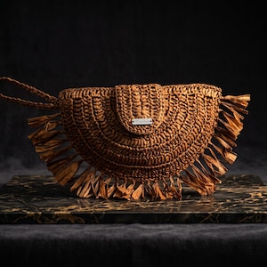 Op de afbeelding: Een halfronde bruine geweven clutch met een textuurrijk vlechtpatroon, een polsbandje aan de linkerkant en lange franjes langs de onderrand. Een klein rechthoekig zilverkleurig metalen label is in het midden op de voorflap geplaatst.