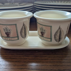 May include: Two off-white ceramic herb pots with a matching tray. Each pot features a framed illustration of a potted plant and a leaf design. The word "sage" is written on the front of each pot. The pots are sitting on a wooden surface.