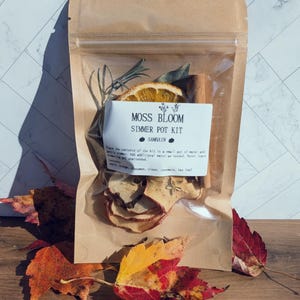 Könnte beinhalten: Ein brauner Papierbeutel mit einem Moss Bloom Simmer Pot Kit für Samhain. Das Sichtfenster zeigt getrocknete Orangenscheiben, Zimtstangen, Apfelscheiben, Nelken, Rosmarin und Lorbeerblätter. Das Etikett enthält Anweisungen und Zutaten.
