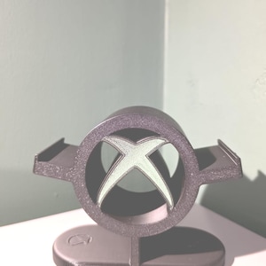 May include: A dark grey 3D-printed controller stand featuring a circular frame with the Xbox logo cutout in the centre. The stand has two side arms for resting a controller and a flat, oval-shaped base with a small logo embossed on the left side.