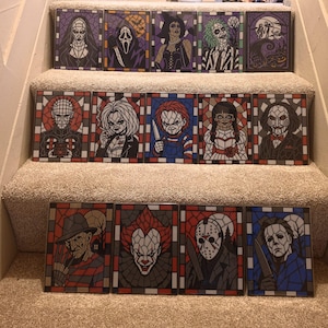 May include: A collection of fourteen rectangular art prints featuring horror movie characters rendered in a stained glass style. Each print displays a distinct iconic figure, including villains and monsters, framed by a geometric border pattern.