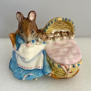 May include: A ceramic figurine of a brown mouse wearing a blue dress and white apron, standing beside a yellow woven basket cradle. Inside the cradle, several small baby mice are tucked under a light pink quilted blanket with blue trim.