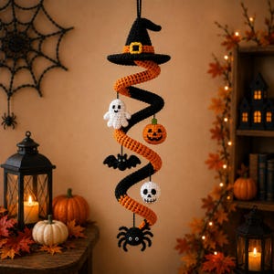Puede incluir: Una decoración de Halloween de ganchillo colgante que presenta un sombrero de bruja negro con una hebilla naranja en la parte superior, unido a una espiral naranja y negra. Pequeñas figuras de ganchillo de un fantasma blanco, una calabaza naranja, un murciélago negro, una calavera blanca y una araña negra cuelgan de la espiral.