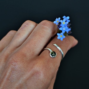 May include: A silver ring with a dark green stone set in a round bezel. The ring is wrapped around the finger and has a simple design. The ring is worn on a hand holding a small bouquet of blue forget-me-not flowers.