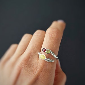 Op de afbeelding: Een zilveren ring met een ontwerp van gele, groene en rode edelstenen. De ring zit aan een vinger.