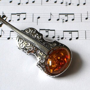 Op de afbeelding: Een zilveren vioolhanger met een inleg van barnsteen. De hanger is gedetailleerd met ingewikkelde krulwerk en ligt op een blad muziek.