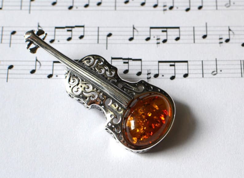 Op de afbeelding: Een zilveren vioolhanger met een inleg van barnsteen. De hanger is gedetailleerd met ingewikkelde krulwerk en ligt op een blad muziek.