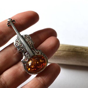 Op de afbeelding: Een zilveren broche in de vorm van een viool met een amberkleurige steen in het midden. De broche is versierd met ingewikkelde uitsnijdingen en wordt in een hand gehouden.