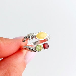 Może przedstawiać: Srebrny pierścionek z żółtym kamieniem w kształcie markizy i dwoma okrągłymi kamieniami, jednym zielonym i jednym czerwonym.