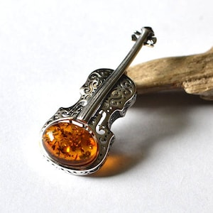 Op de afbeelding: Een zilveren viool broche met een amber steen in het midden. De viool is gedetailleerd met ingewikkelde gravures en de amber steen heeft een rijke, warme kleur.