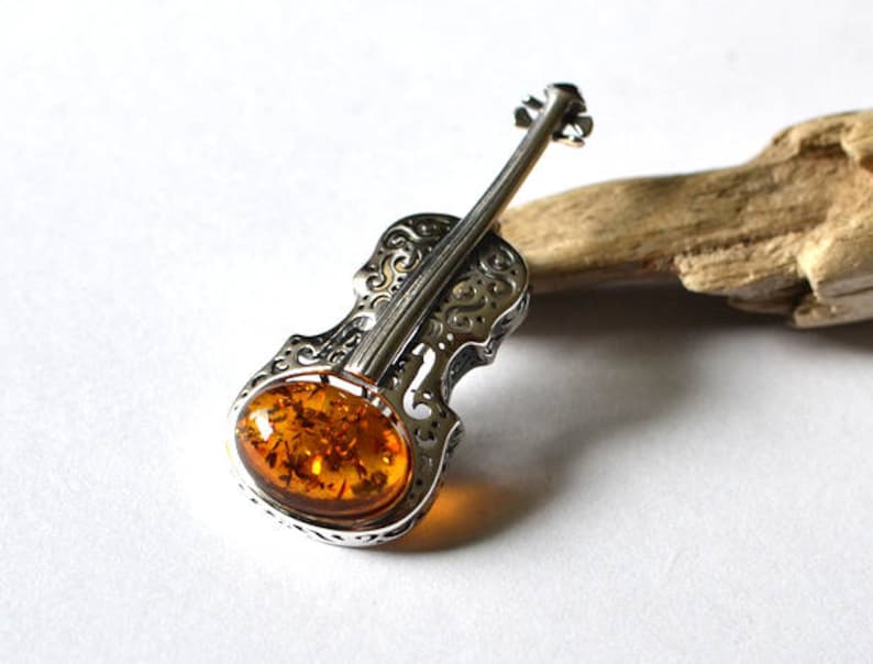 Op de afbeelding: Een zilveren viool broche met een amber steen in het midden. De viool is gedetailleerd met ingewikkelde gravures en de amber steen heeft een rijke, warme kleur.