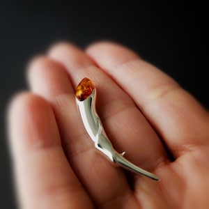 Op de afbeelding: Zilverkleurige broche met een gepolijste afwerking, met een druppelvormige barnsteen aan de bovenkant. De broche heeft een gebogen, organisch ontwerp met een puntig uiteinde. De barnsteen heeft een warme, goudoranje kleur.