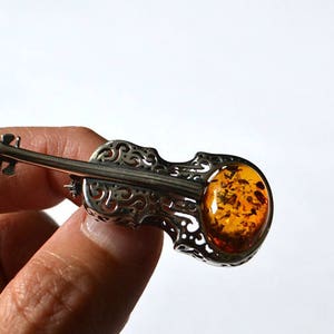 Op de afbeelding: Een zilveren broche in de vorm van een viool met een amberkleurige steen in het midden.