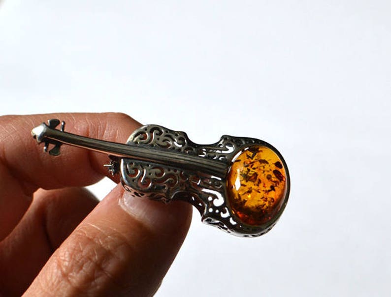Op de afbeelding: Een zilveren broche in de vorm van een viool met een amberkleurige steen in het midden.
