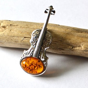 Op de afbeelding: Een zilveren viool broche met een amber steen in het midden. De viool is versierd met ingewikkelde krulwerk.