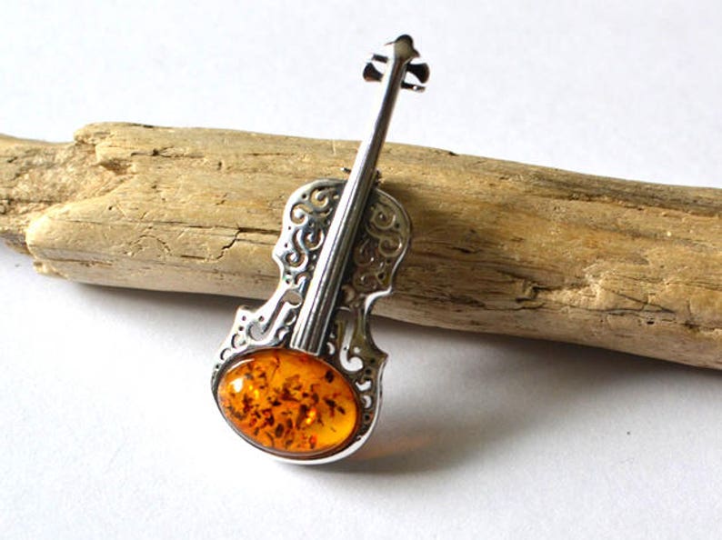 Op de afbeelding: Een zilveren viool broche met een amber steen in het midden. De viool is versierd met ingewikkelde krulwerk.