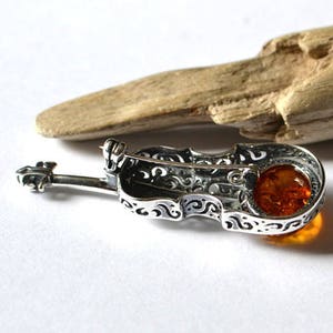 Op de afbeelding: Zilveren viool broche met een amber steen in het midden. De viool is versierd met ingewikkelde krulwerk.