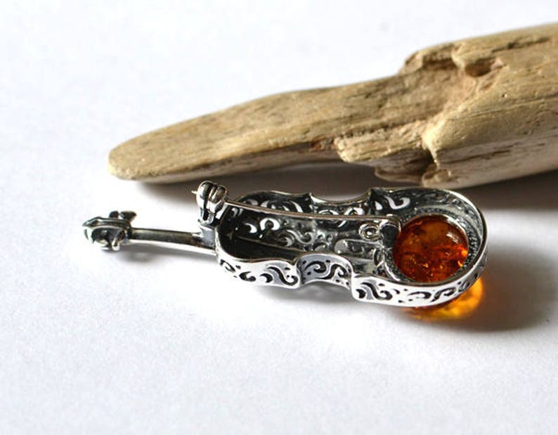 Op de afbeelding: Zilveren viool broche met een amber steen in het midden. De viool is versierd met ingewikkelde krulwerk.