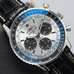 May include: A Breitling Navitimer chronograph watch with a silver dial, three black sub-dials, and a blue slide rule bezel. The watch features a stainless steel case, a date window, and a black leather strap with a textured crocodile pattern.