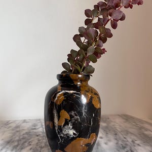May include: A polished stone vase featuring a dark black base with prominent patches of deep amber and white veining. The vase has a rounded body and a short, narrow neck. A single stem with small, dark reddish-purple leaves is placed inside.