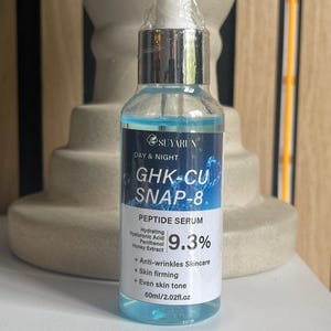 以下が含まれることがあります： 透明なガラス製のスポイトボトルに入った青い液体セラム。ラベルには「Suyarun Day & Night GHK-CU SNAP-8 Peptide Serum 9.3%」と記載され、ヒアルロン酸、パンテノール、ハチミツエキスが含まれています。シワ防止、肌の引き締め、肌のトーンを整える効果に関するテキストがあり、容量は60mlです。