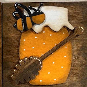 May include: Wooden honey jar with a bee and honey dipper. The jar is orange with white polka dots, topped with a white lid. A black and yellow bee is perched above the jar. The honey dipper is brown.