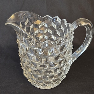 Puede incluir: Una jarra de cristal transparente que presenta un patrón geométrico de cubos en toda su superficie exterior. El recipiente incluye un asa curva en el lateral y un pico vertedor ancho y moldeado en el borde superior, con diseño facetado.