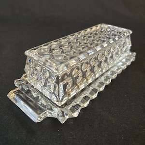 May include: A clear pressed glass butter dish featuring a rectangular base with a scalloped edge and a matching lid. The lid is decorated with a raised diamond pattern across its top and sides, creating a textured, faceted appearance throughout.