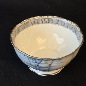 May include: A white porcelain bowl featuring a hand-painted landscape scene with birch trees on the exterior. The rim is decorated with a grey and white textured pattern and finished with a thin gold trim. The base also features gold detailing.