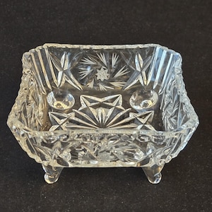 May include: A clear, square-shaped crystal bowl featuring intricate geometric starburst patterns cut into the glass. The bowl is supported by four small, sturdy feet and has a faceted rim, showcasing a reflective and transparent design.