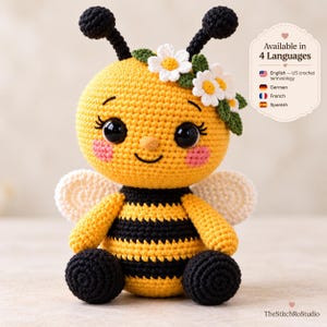 May include: A crocheted bee plush toy with a yellow and black striped body, large black eyes, pink cheeks, and white wings. It features a crown of white flowers with green leaves on its head and two black antennae. Text reads Available in 4 Languages.
