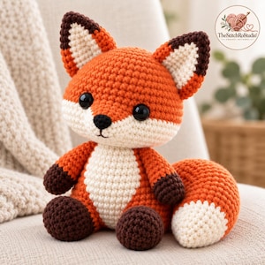 Può includere: Un peluche a forma di volpe realizzato all'uncinetto con pelliccia arancione brillante, petto e muso color crema, zampe marrone scuro e orecchie a punta con centro color crema. La volpe ha grandi occhi neri di sicurezza e un piccolo naso ricamato nero. Il logo StitchRoStudio è visibile.