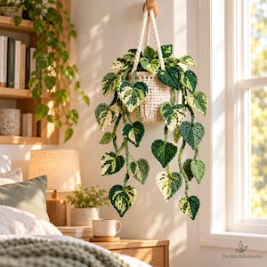 Op de afbeelding: Een gehaakte hangplant in een crèmekleurige pot, met bonte hartvormige bladeren in tinten donkergroen, lichtgroen en crème. De plant hangt aan een bijpassend gehaakt koord en is voorzien van de tekst TheStitchRoStudio.