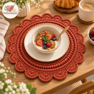 以下が含まれることがあります： スカラップエッジが特徴の錆色の円形かぎ針編みプレースマット。質感のある円形のステッチパターンが施されています。プレースマットの中央には、ヨーグルト、グラノーラ、スライスしたイチゴ、ブルーベリーが入った白いセラミック製のボウルが置かれています。