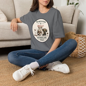 May include: A dark gray t-shirt featuring a circular graphic with a cartoon squirrel holding acorns and papers. The text reads, The squirrel brain is REALLY SQUIRRELING TODAY. The design includes small acorn illustrations surrounding the central squirrel figure.